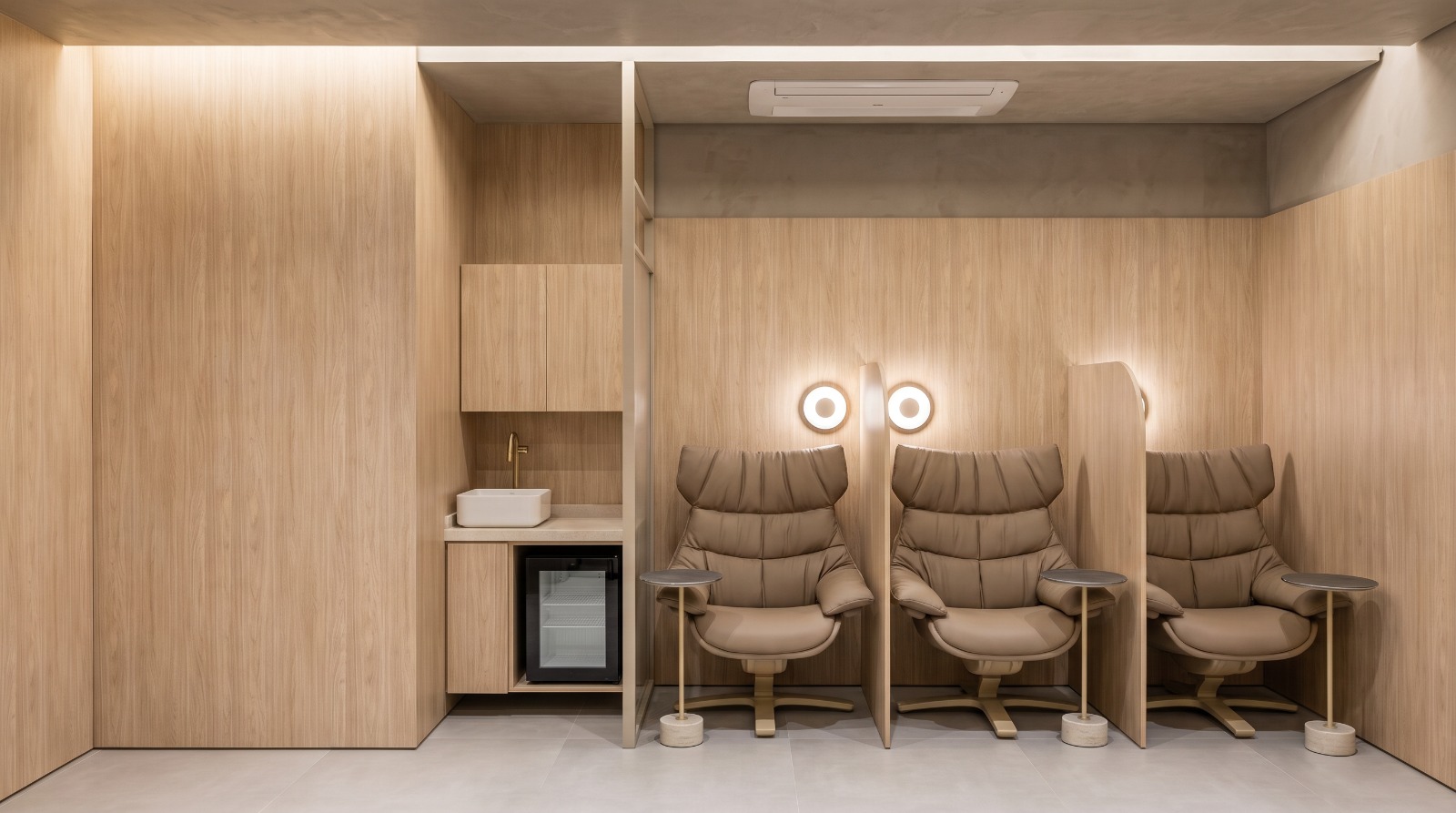 Sala de infusão da clínica com poltronas em couro taupe, divisórias de madeira clara e iluminação suave